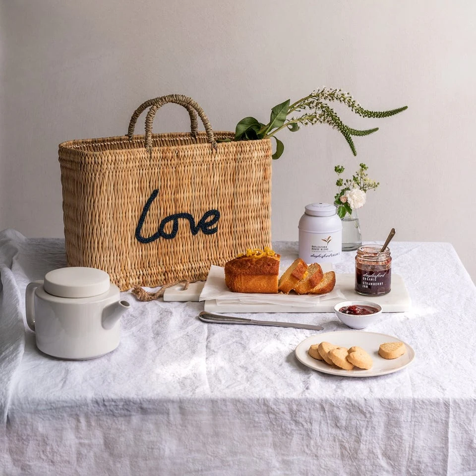 Woven Love basket bag, tea, jam, cake and biscuits arranged on a linen-covered table