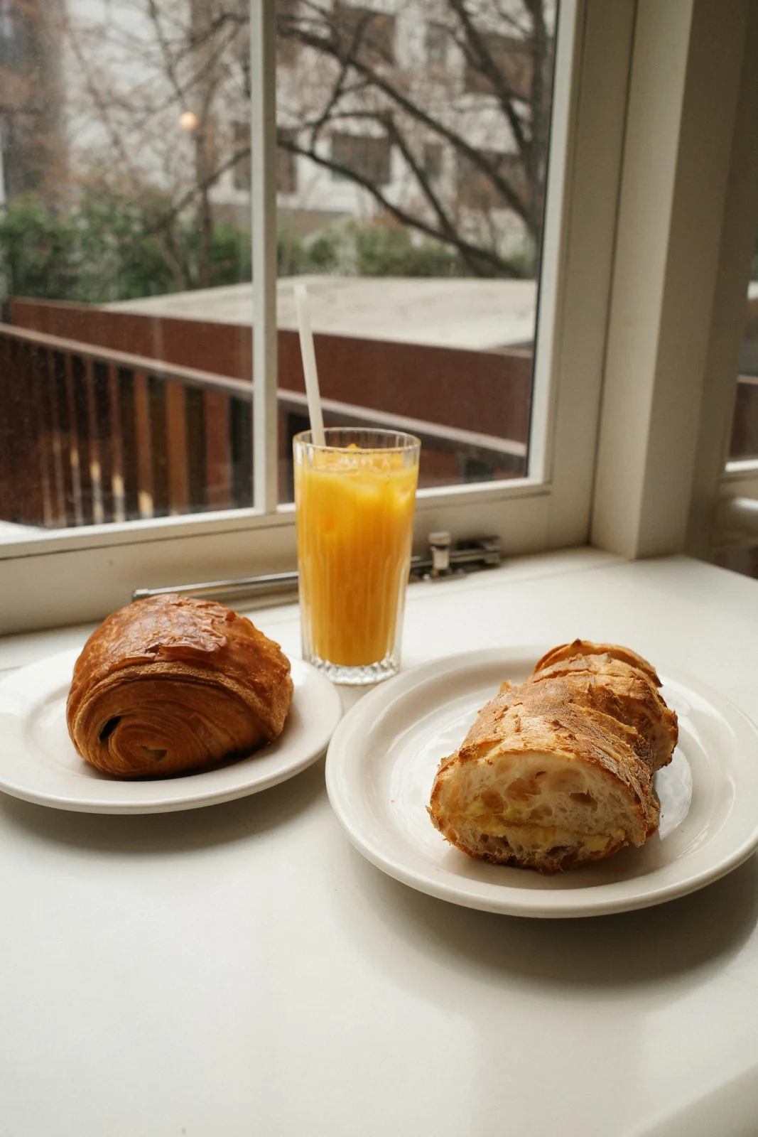 Two croissants on white plates and a glass of orange juice on a windowsill.