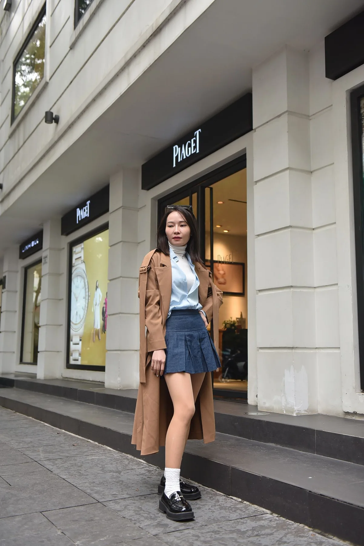 Woman wearing a long brown coat over a short pleated skirt on a shopping street.