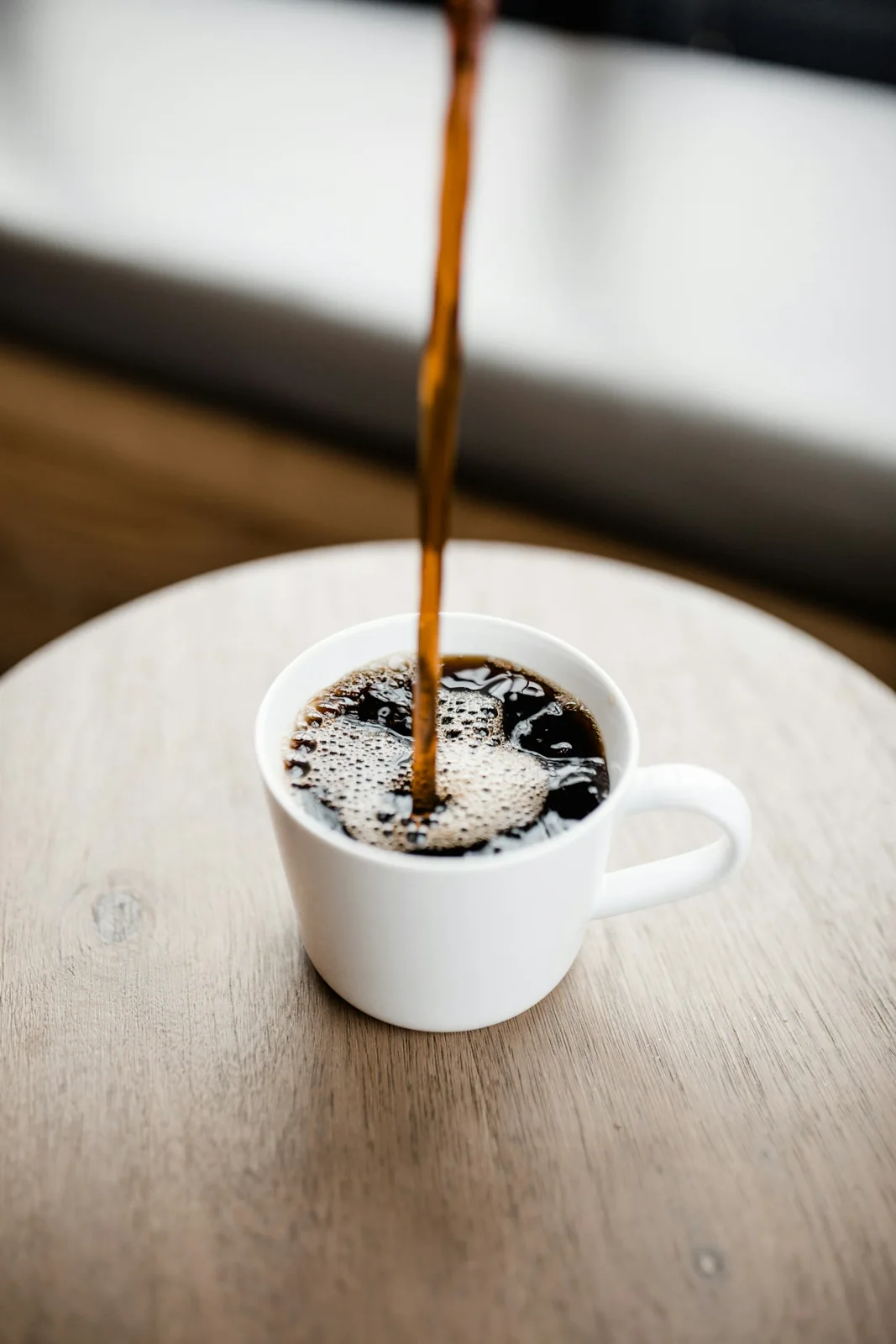 Dark coffee being poured into a white ceramic cup on a wooden tabletop.