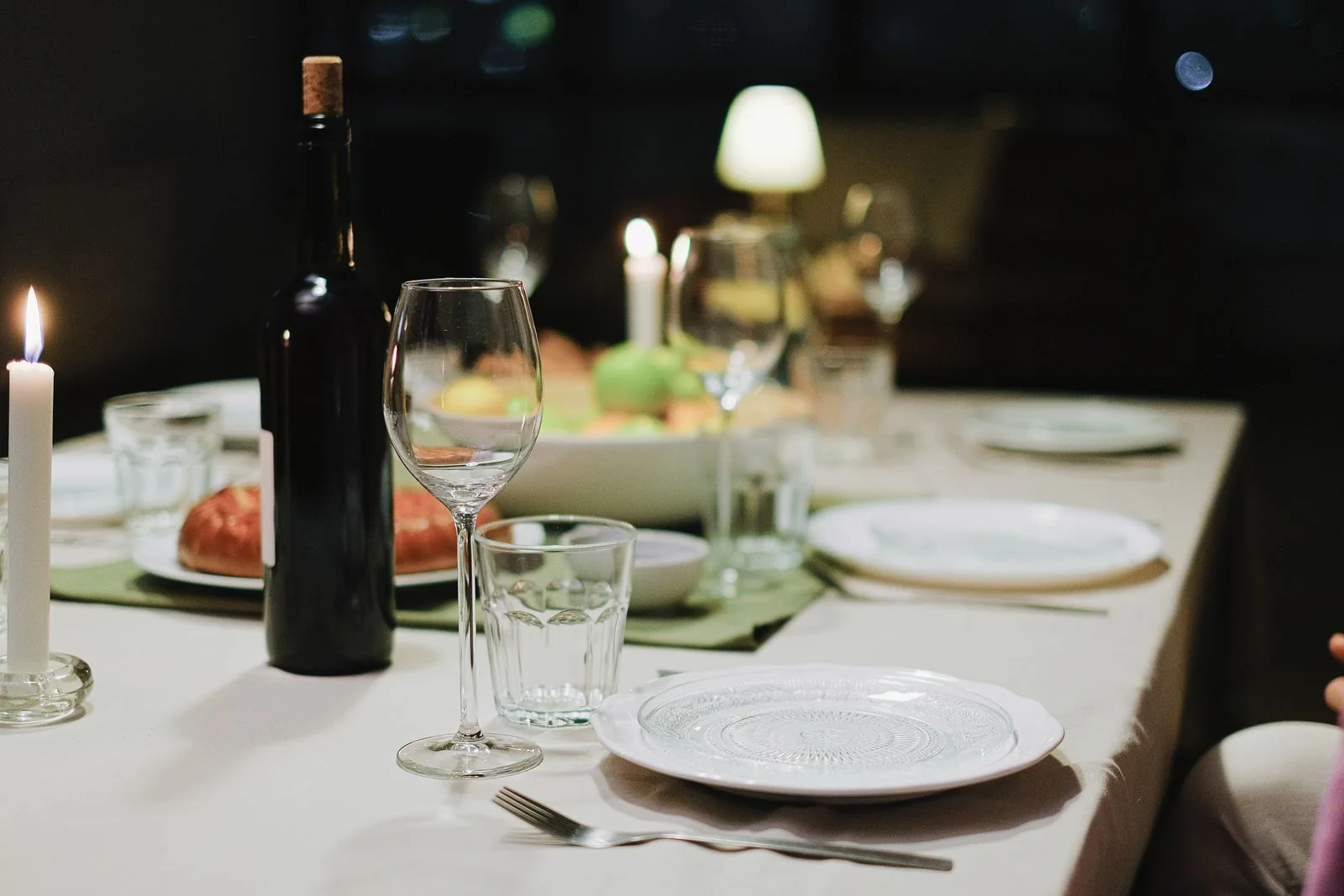 Dinner table set with white plates, cutlery, glasses and a dark bottle.