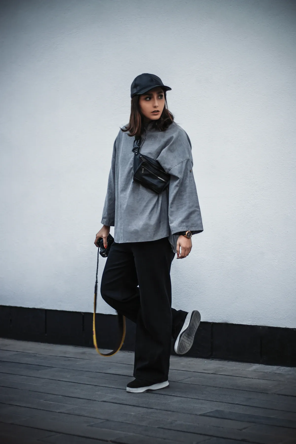 Woman wearing a gray top, dark wide-leg trousers and a black cap against a light wall.