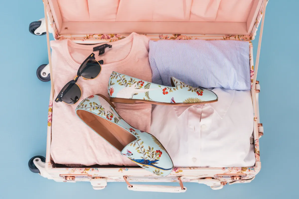 Open pink suitcase containing clothing, shoes and sunglasses.