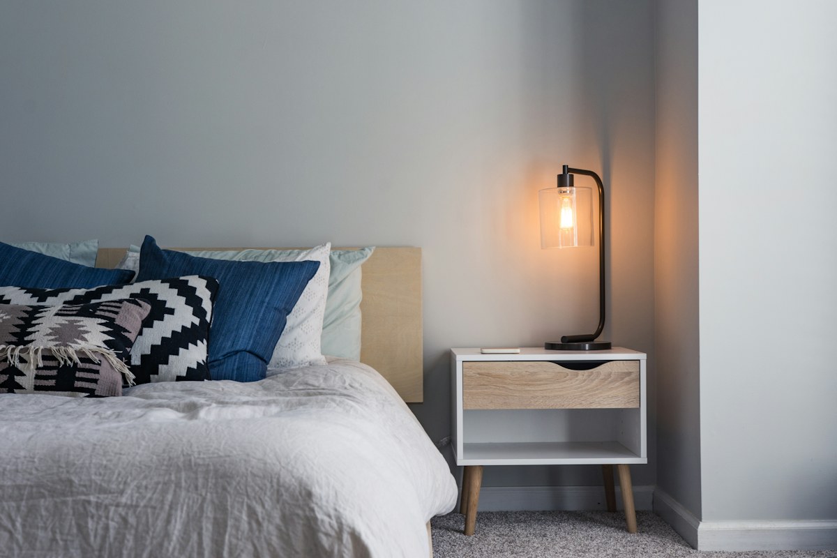 A slim black bedside lamp beside a bed with blue cushions.