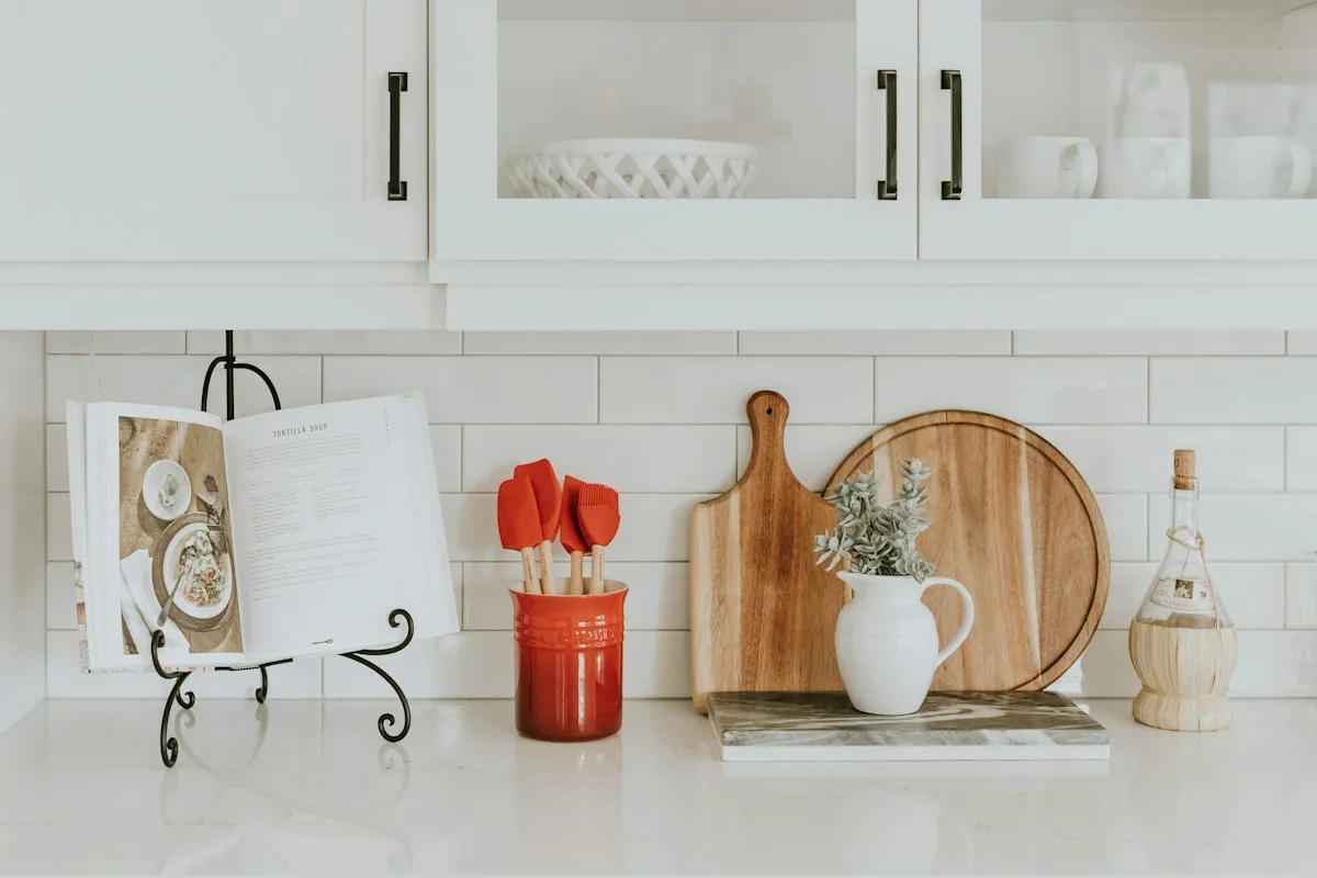 A pale kitchen counter with wooden boards, red utensils and a white jug.