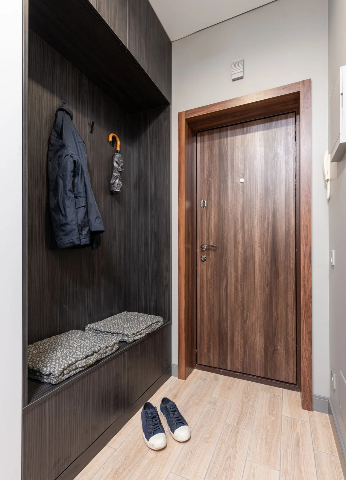 A compact entryway with a wooden door, hanging coats and a cushioned bench.