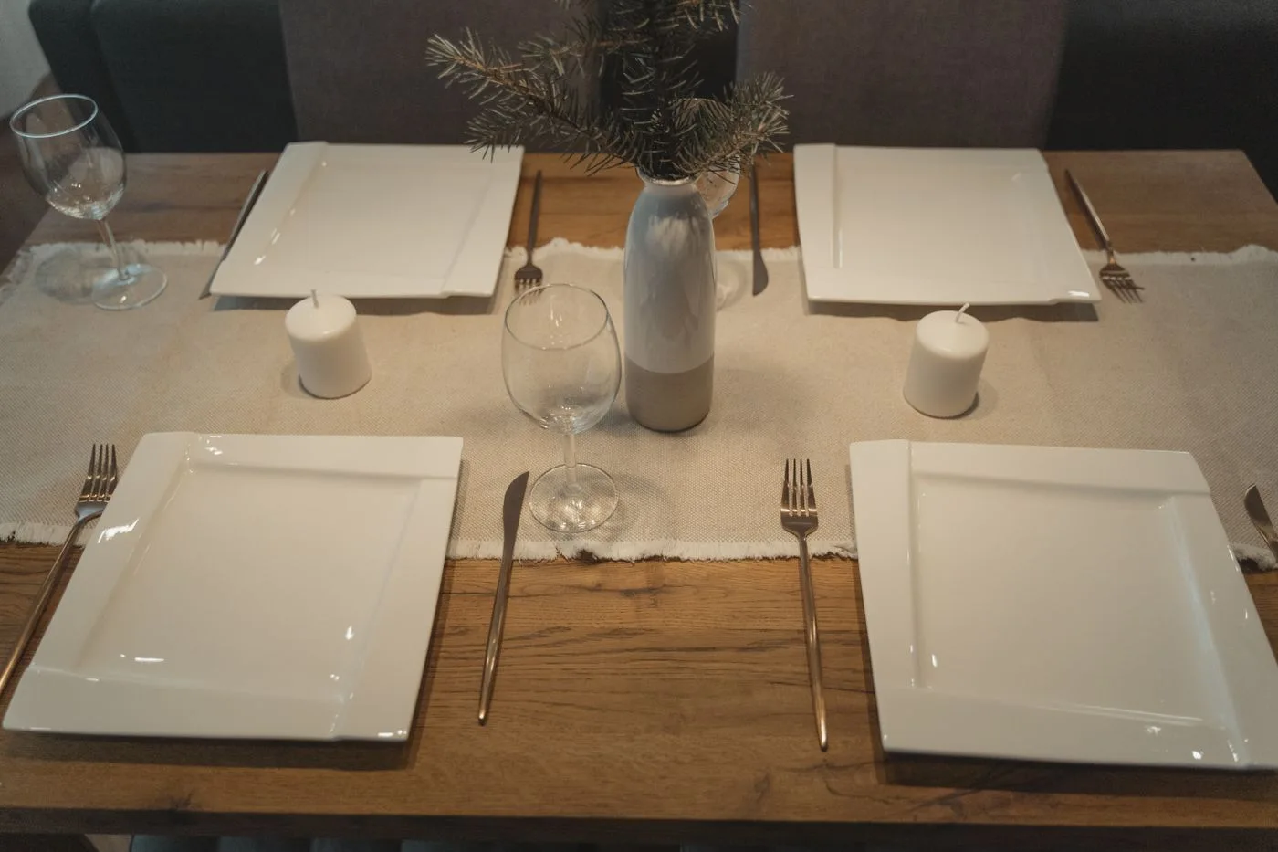 A wooden dining table laid with square white plates, glasses and small cups.