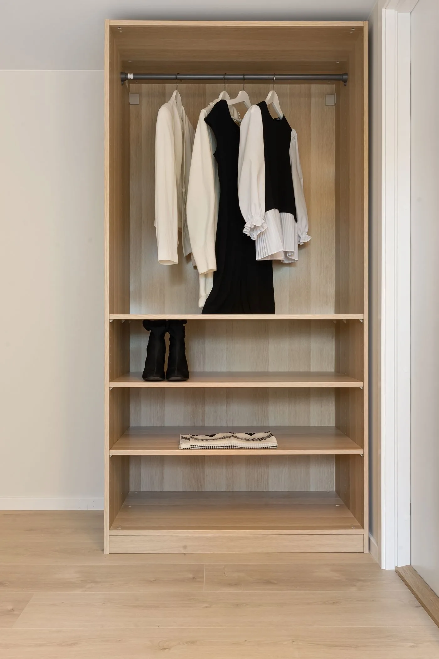 An open wooden wardrobe with hanging clothes and several mostly empty shelves.