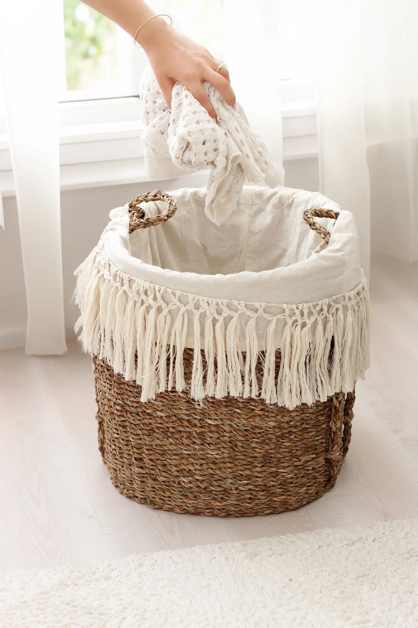 A hand lowering pale laundry into a fringed wicker basket beside a window.