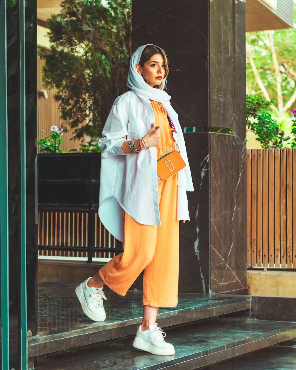 Woman wearing a pale blue open shirt over an orange outfit with white sneakers outdoors.