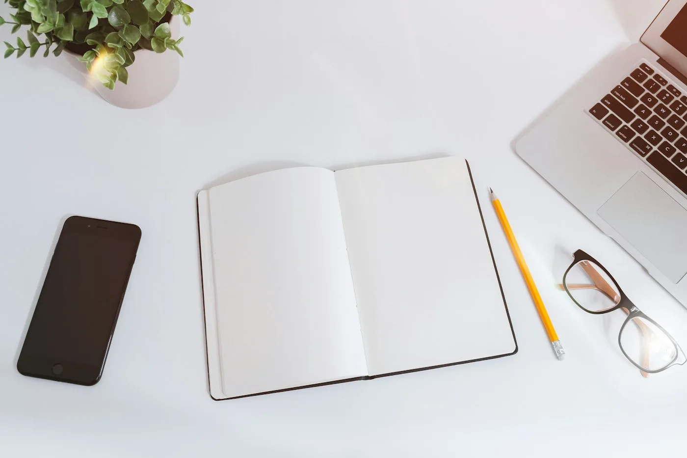 An open notebook with writing tools, glasses and devices on a white desk.