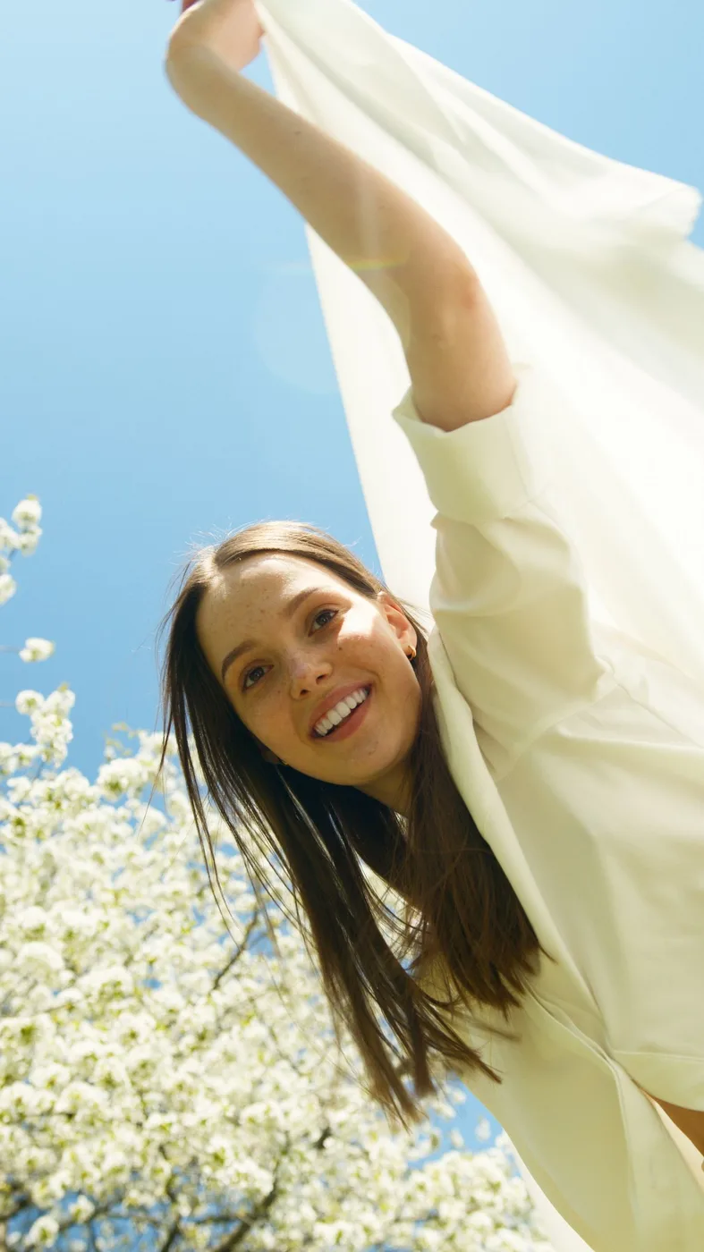 A woman wearing a loose light top outdoors with one arm raised.